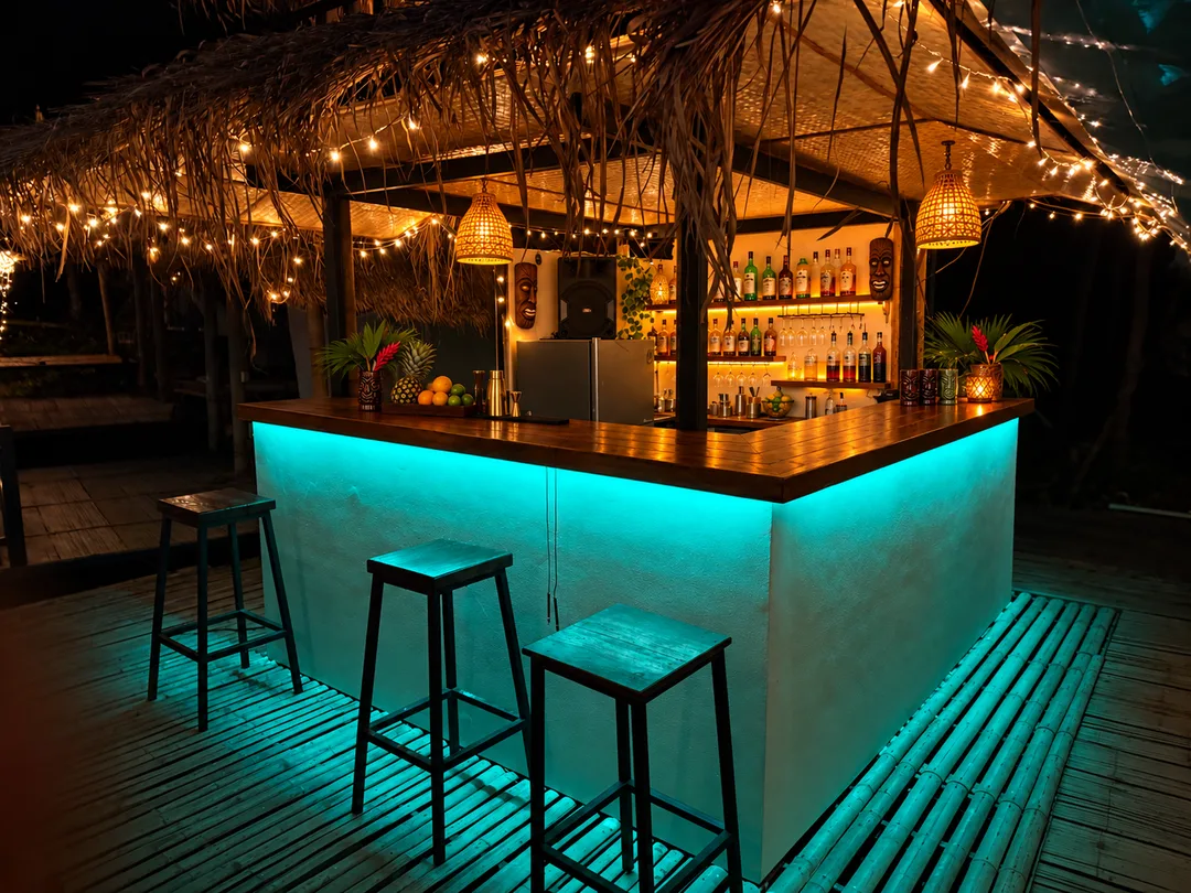 Turquoise-lit bar counter, wooden stools and string lights at Oasis Tiki Bar at night.